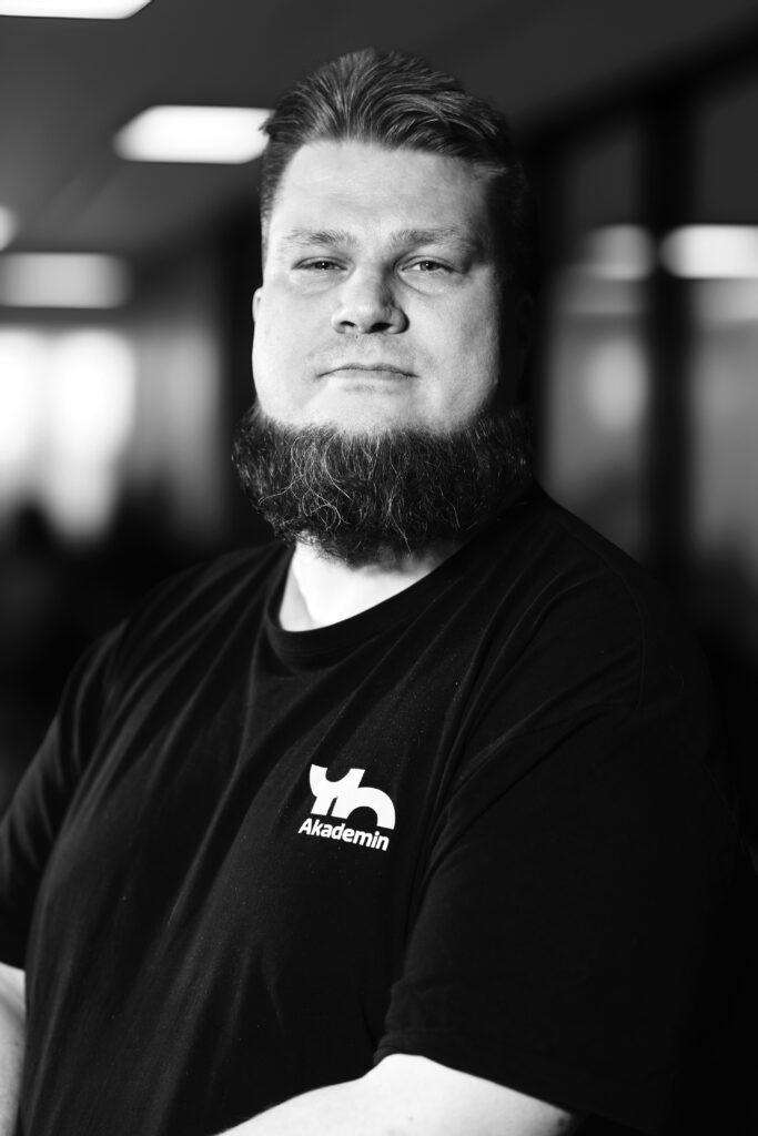 En skäggig person i svart skjorta står självsäkert inomhus.