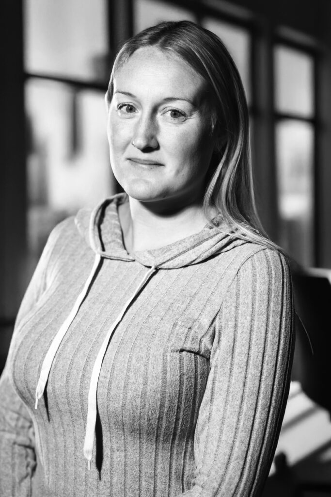 Kvinna med långt hår i randig luvtröja tittar på kameran, står inomhus med stora fönster i bakgrunden.
