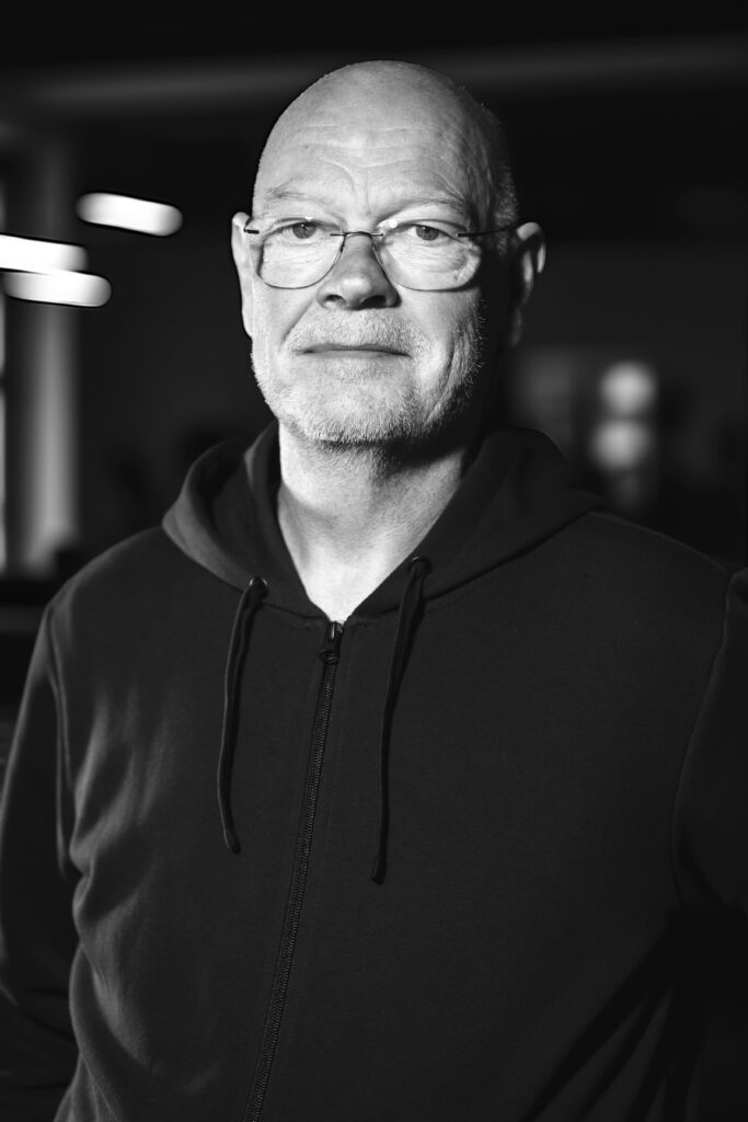 Äldre man med glasögon och skägg i luvtröja, stående inomhus med svag belysning. Svartvitt foto.