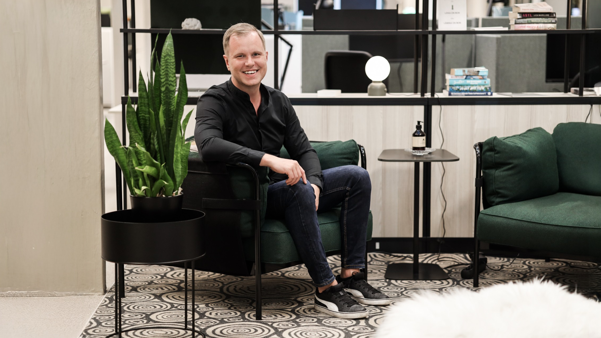 En leende man i svart skjorta sitter på en grön stol bredvid en växt i ett modernt kontorsrum.