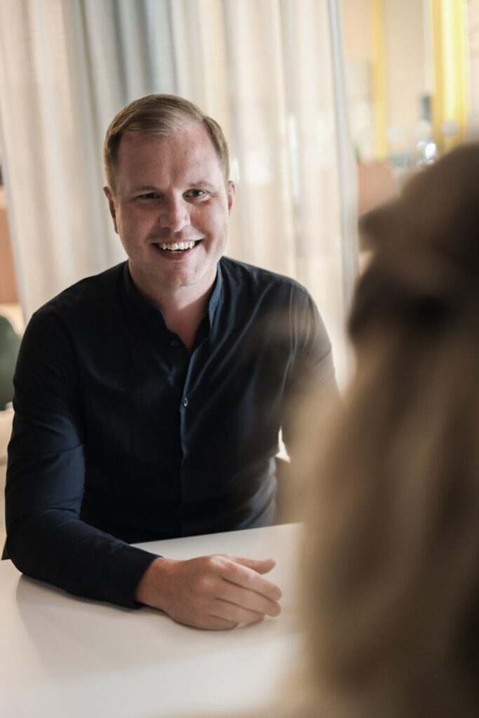 En leende affärsutvecklare besöksnäringen i mörk skjorta sitter vid ett bord och pratar med någon som har ryggen mot kameran.