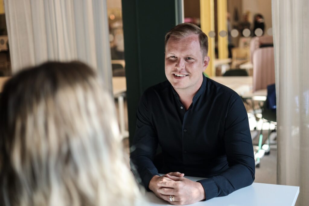 En man i mörk skjorta ler och pratar med en kvinna tvärs över ett bord i ett ljust rum.