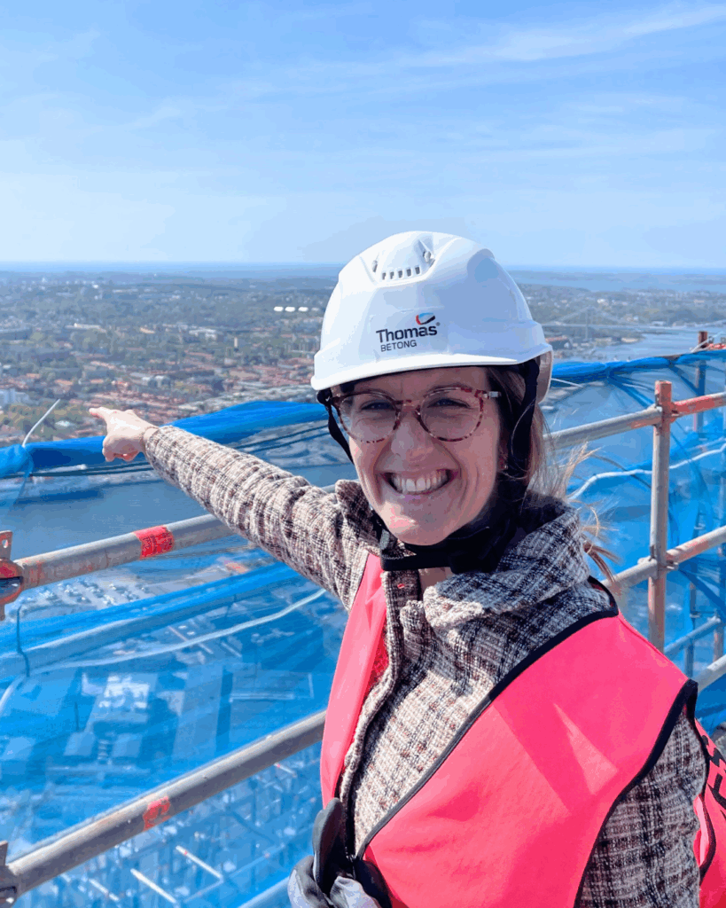 Leende Betongingenjör i skyddsutrustning pekar ut över en stad från en hög byggnadsställning.