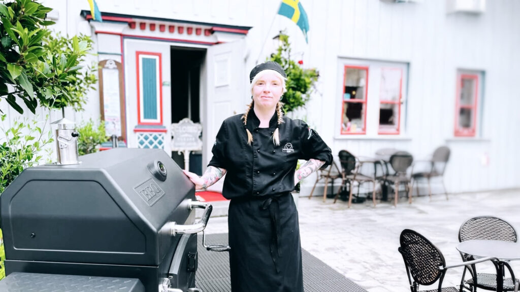 En gastronomiker står vid en grill utanför en restaurang med uteplats med sittplatser och färgglad dekoration.