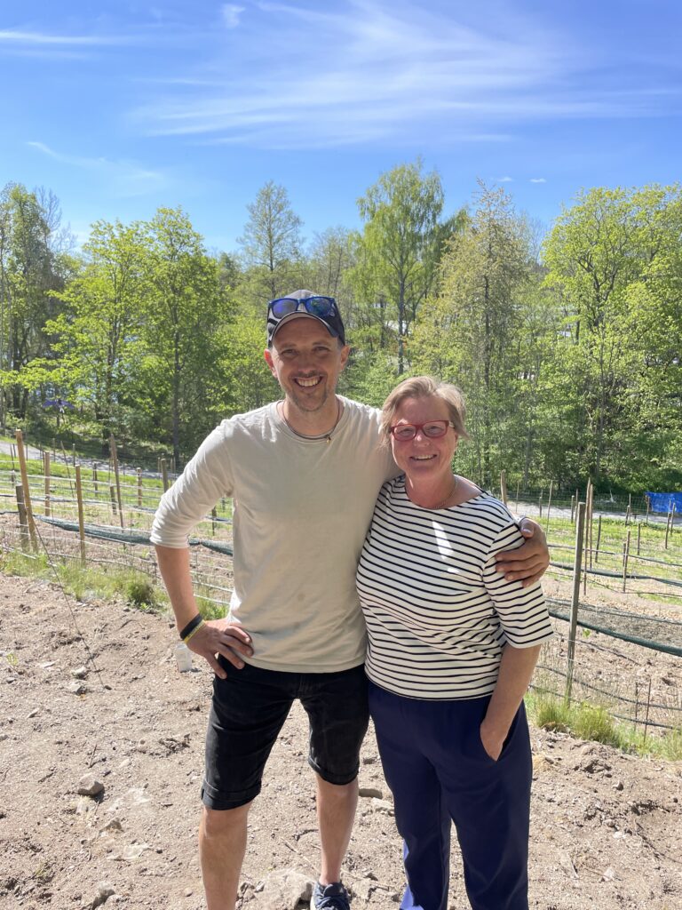 Två personer ler utomhus, står arm i arm på en solig dag nära en vingård med träd i bakgrunden.
