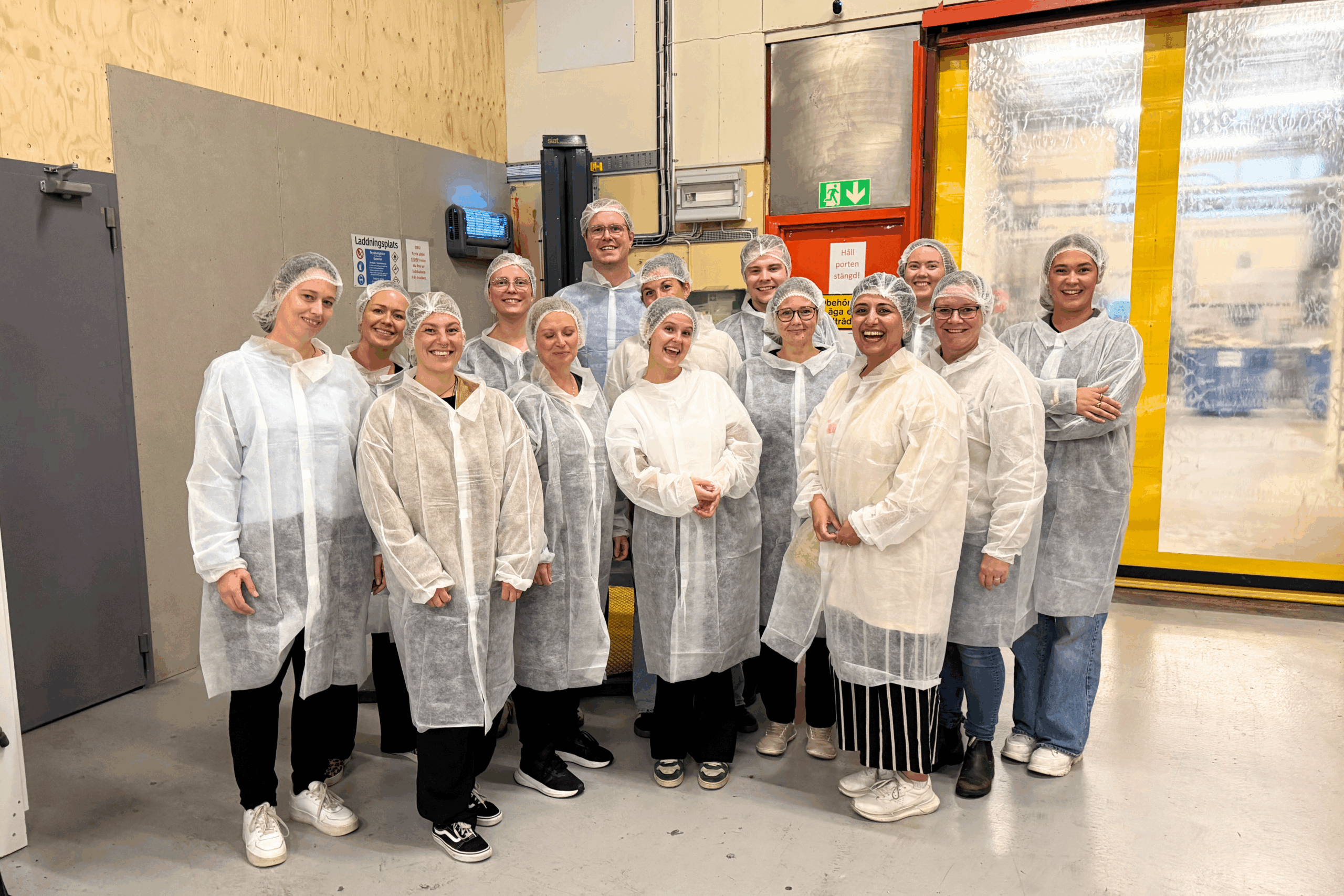 En grupp människor i labbrockar, hårnät och skoskydd ler inne i en industrianläggning, redo för sin Terminsstart.