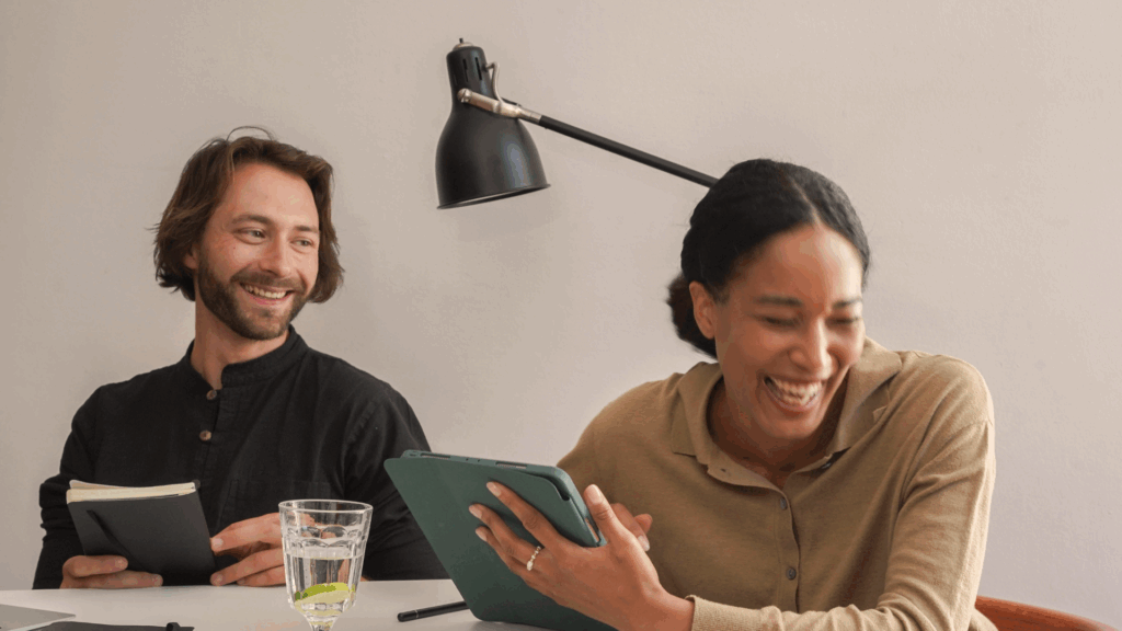 Två personer sitter vid ett bord och skrattar tillsammans; den ena håller i en surfplatta, den andra i en anteckningsbok.
