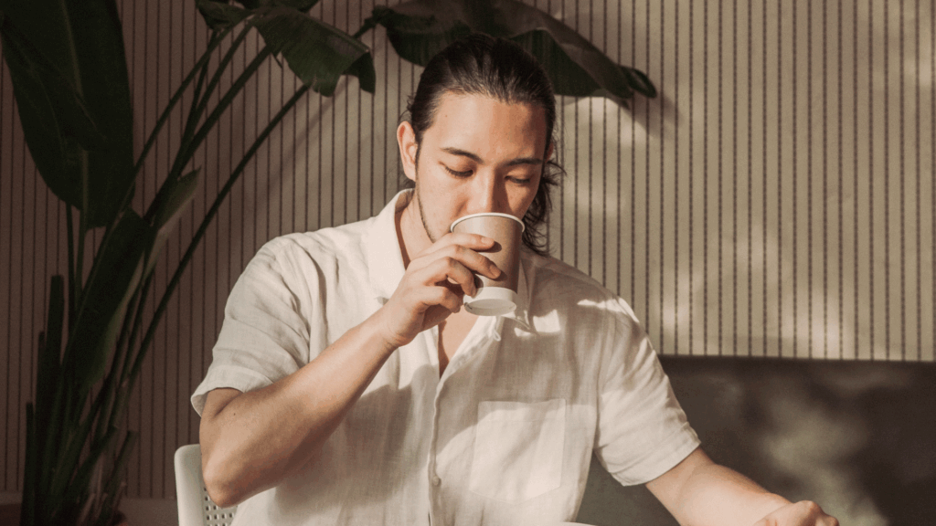 Man med långt hår i vit skjorta som dricker ur en pappersmugg, sittande inomhus med växter i bakgrunden.