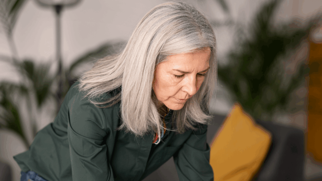 Äldre kvinna med grått hår och grön skjorta tittar ner, fokuserad, i ett mysigt rum med växter och en gul kudde - kanske arbetar hon med sin sen anmälan.