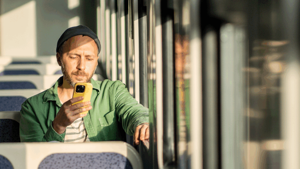 En man i grön skjorta och hatt sitter ensam på ett solbelyst tåg, fokuserad på sin telefon medan han fyller i en ansökan.