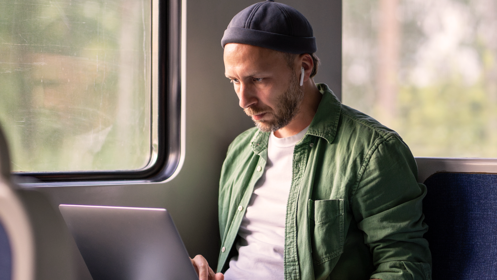 Man i grön tröja och mössa arbetar på laptop medan han åker tåg, med trådlösa öronsnäckor, fokuserad på att slutföra en viktig ansökan.