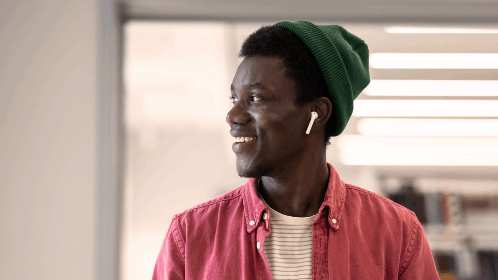 Leende ung man i grön mössa och hörlurar, klädd i röd skjorta, tittar åt sidan inomhus - kanske tänker han på sin ansökan.