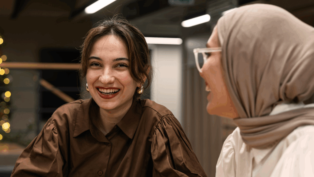 Två kvinnor ler och pratar inomhus, den ena bär en brun skjorta, den andra en beige hijab och glasögon.