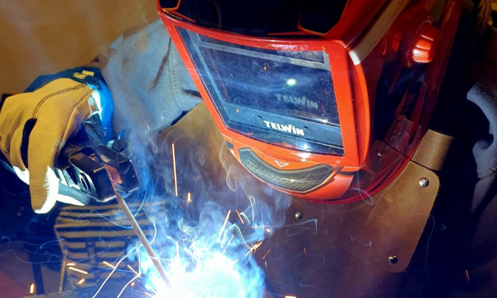 En person med svetshjälm och handskar svetsar försiktigt metall, med starka gnistor och blått ljus som belyser scenen - förbereder sig för nästa Terminsstart.