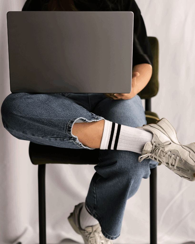 Person som sitter med benen i kors på en stol och använder en bärbar dator, klädd i jeans, vita strumpor och sneakers.