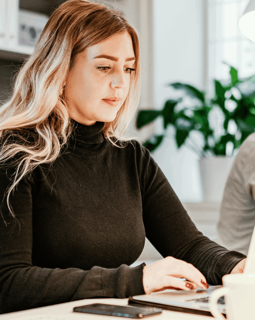 Kvinna med långt blont hår sitter vid ett skrivbord och pluggar till sin preparandkurs, med en telefon och kaffekopp i närheten.