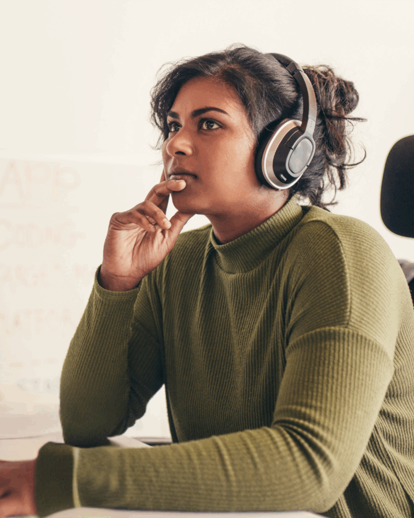 Kvinna i grön tröja och hörlurar sitter vid sitt skrivbord och ser eftertänksam ut med handen på hakan - kanske funderar hon på nästa steg i en förberedelsekurs.