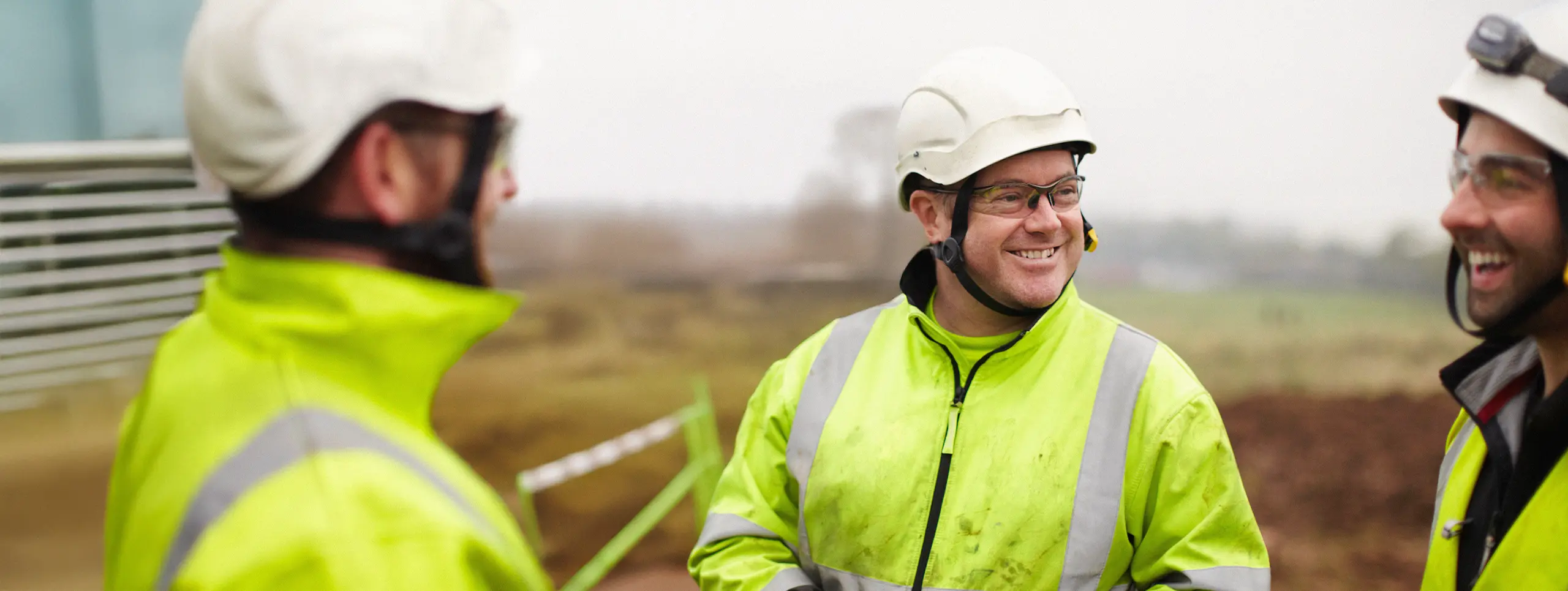 Tre byggnadsarbetare i varseljackor och hjälmar ler och pratar på en arbetsplats utomhus.