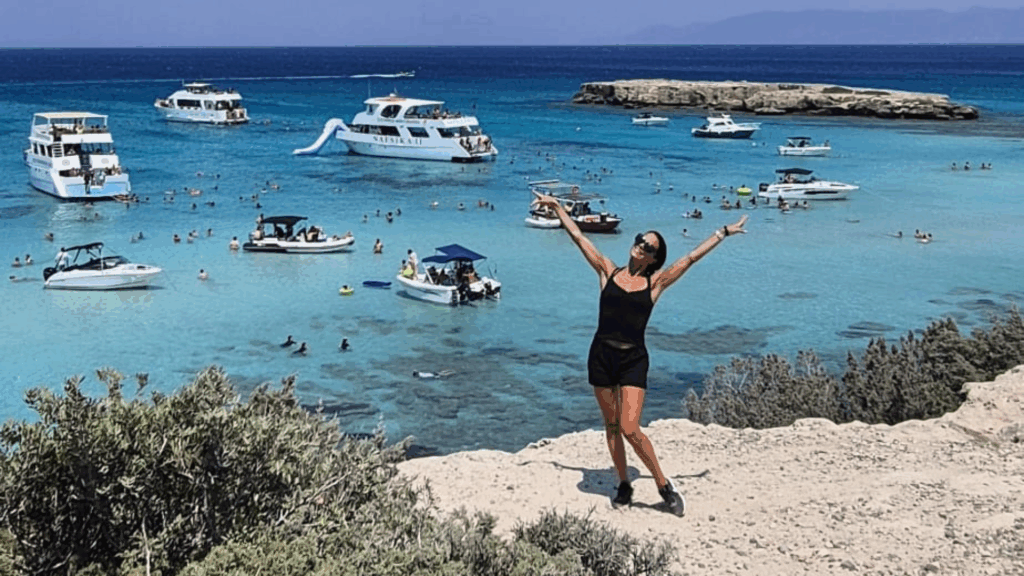 En kvinna poserar glatt på en klippa med utsikt över det turkosa havet med båtar och simmare i bakgrunden - ett exempel på Betongingenjörens äventyrsanda som omfamnar naturen.
