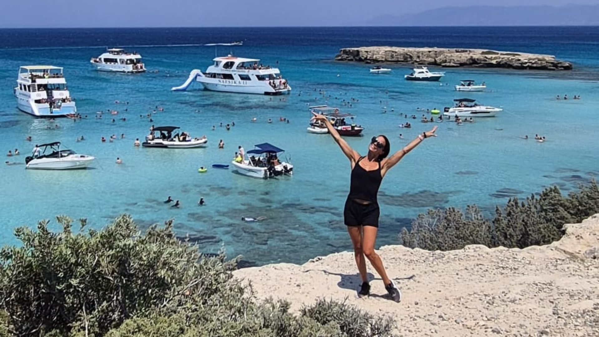 En kvinna poserar glatt på en klippa med utsikt över det turkosa havet med båtar och simmare i bakgrunden - ett exempel på Betongingenjörens äventyrsanda som omfamnar naturen.