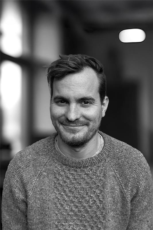 En man med kort hår och skägg, klädd i stickad tröja, ler mot kameran inomhus.
