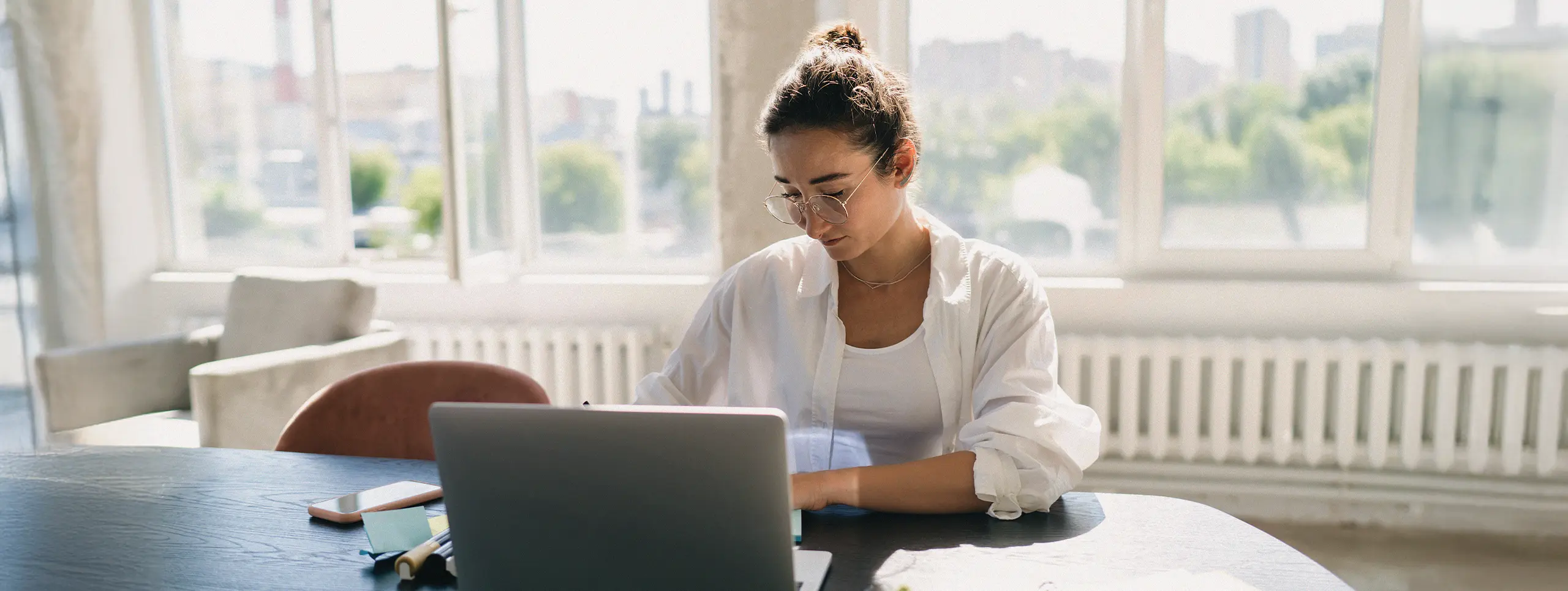 Kvinna med glasögon arbetar vid en bärbar dator i ett ljust rum med anteckningsböcker och papper på bordet.