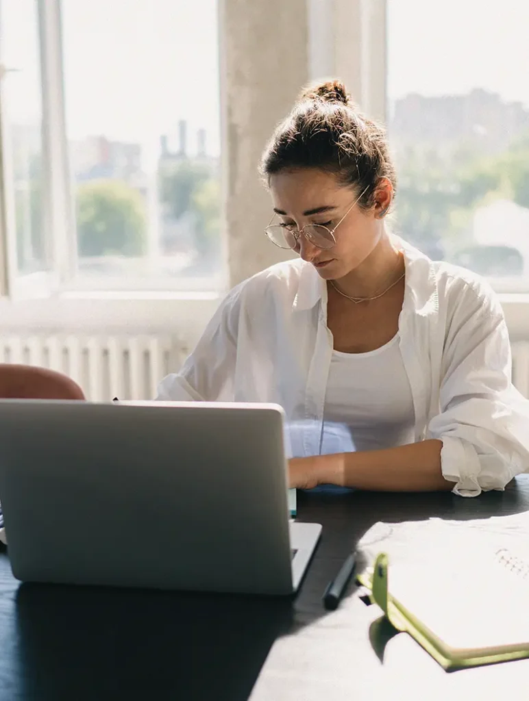 Kvinna med glasögon arbetar på en bärbar dator vid ett skrivbord nära ett ljust fönster, med anteckningsböcker och kontorsmaterial i närheten.