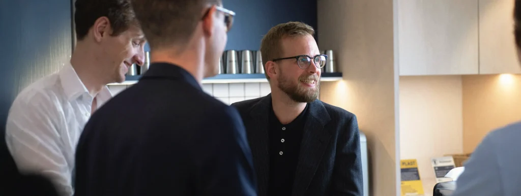 Fyra personer i affärskläder pratar och ler runt ett träbord i ett modernt kontorskök.