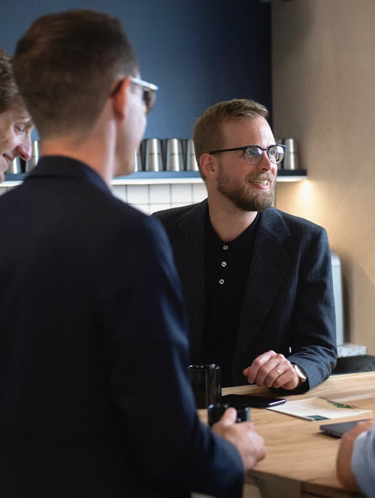 Tre personer i affärsdräkt diskuterar runt ett bord i en modern kontorsmiljö.