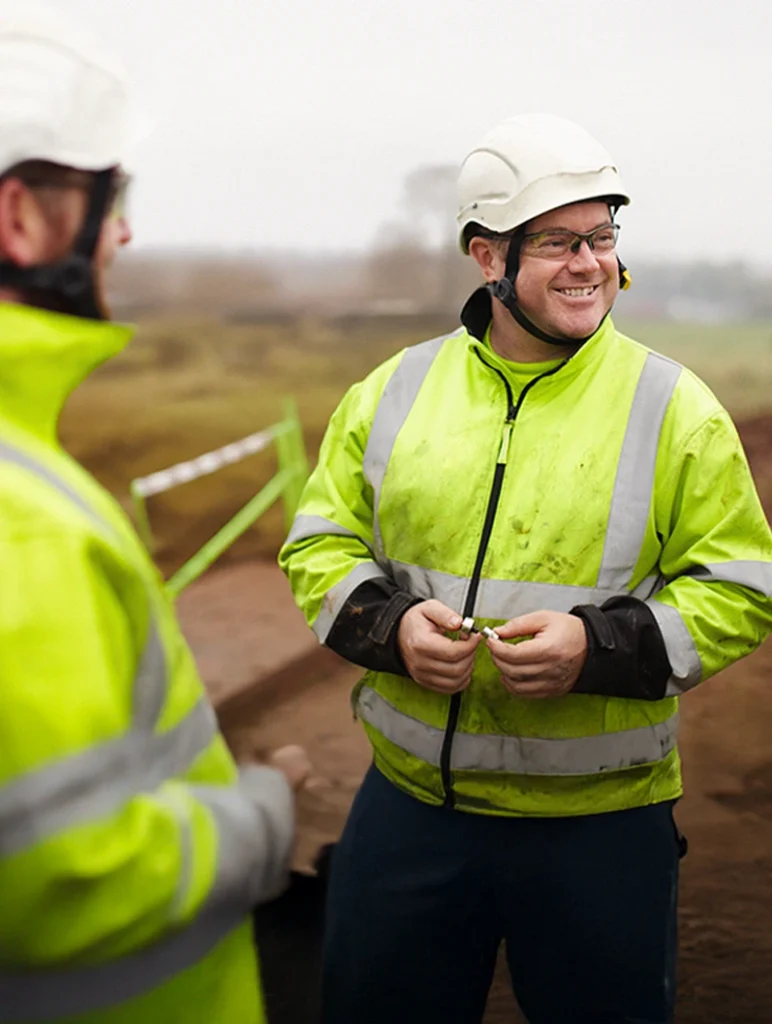 Tre byggnadsarbetare i gula skyddsvästar och hjälmar ler och pratar på en arbetsplats utomhus.