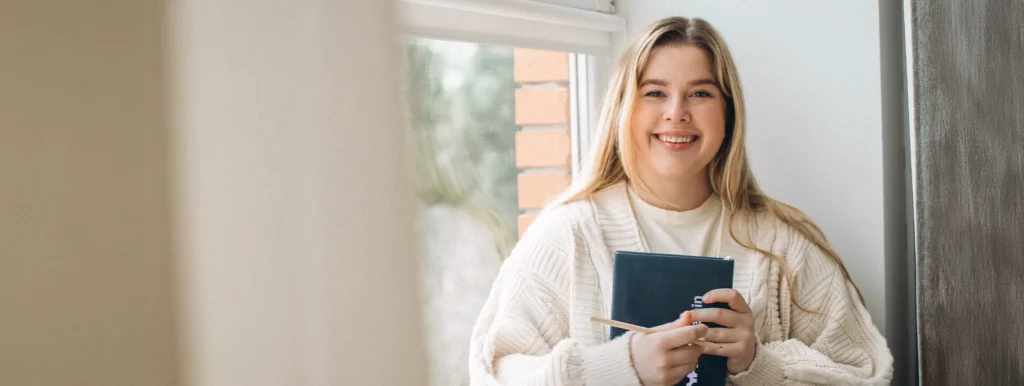 Leende kvinna i gräddfärgad tröja sitter vid ett fönster och håller i anteckningsbok och penna.