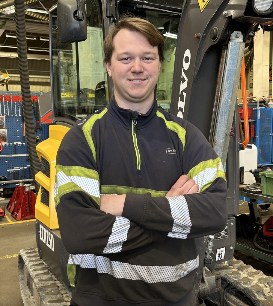 En elingenjör i reflekterande arbetsjacka står med armarna i kors framför en byggutrustning i en verkstad.