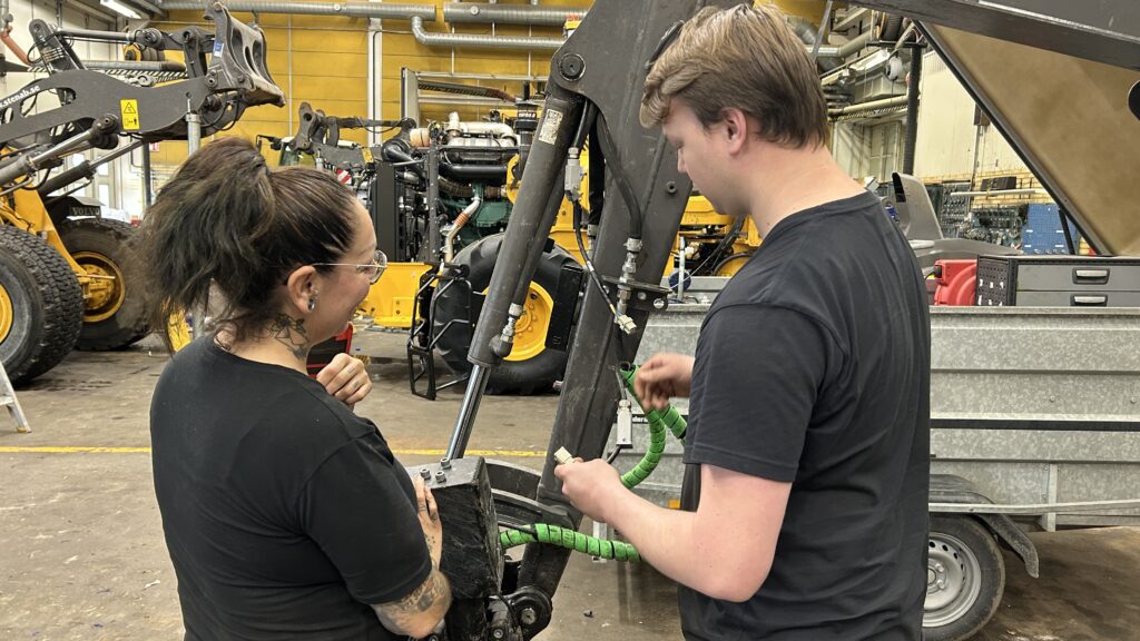 Två personer i en verkstad, inklusive en elingenjör, undersöker och diskuterar maskiner, med verktyg och utrustning synliga i bakgrunden.