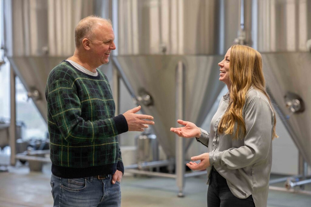 Två personer ler och pratar framför stora metalltankar i ett bryggeri.
