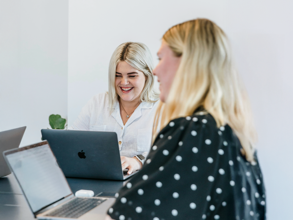 Två kvinnor med bärbara datorer samarbetar vid ett bord, den ena ler, i en ljus, modern kontorsmiljö på en yrkeshögskola.