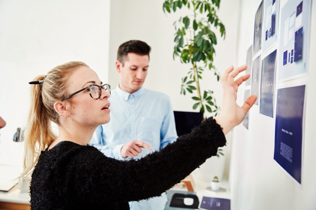 Kvinna med glasögon pekar på designlayouter på en vägg medan en man står i närheten och observerar - ett kreativt ögonblick på YH Akademin.