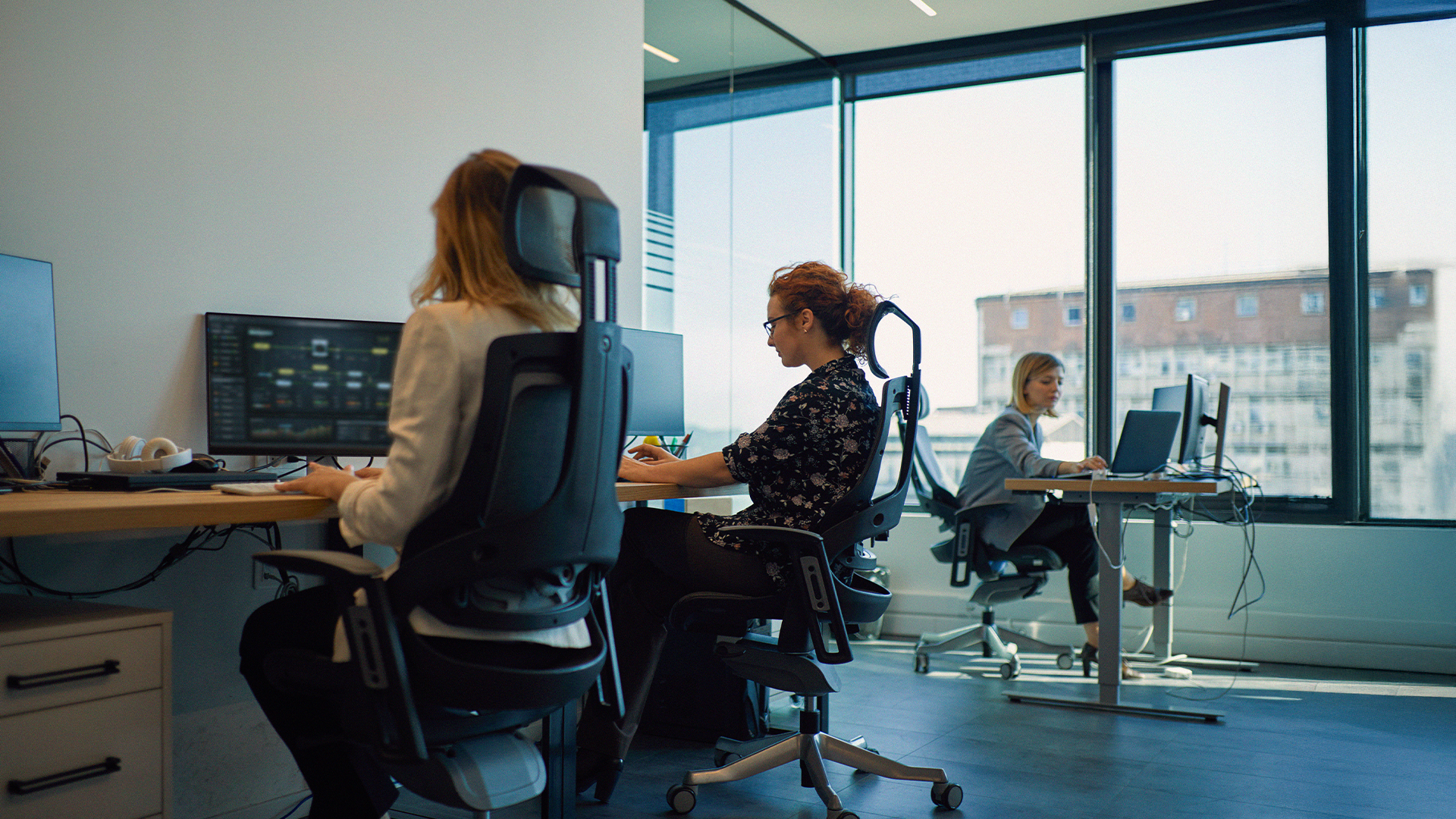 Tre kvinnor arbetar vid skrivbord med datorer i ett modernt kontor med stora fönster och naturligt ljusinsläpp.