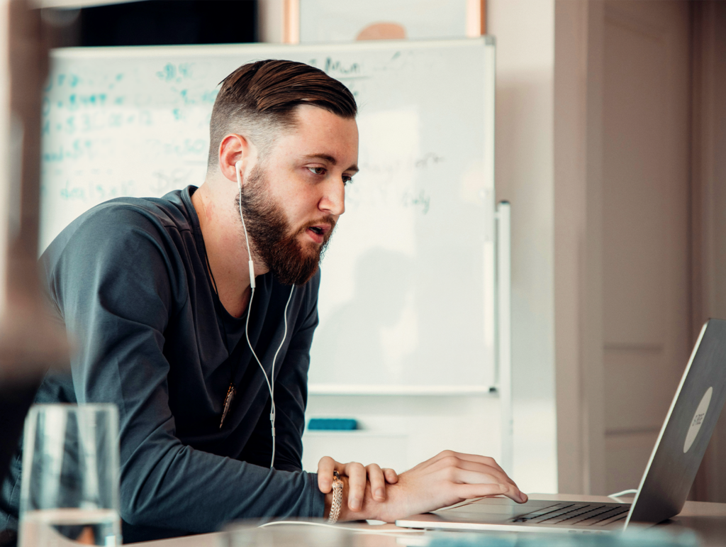 Man med hörlurar som arbetar på en bärbar dator vid ett skrivbord, whiteboard med skrift i bakgrunden - perfekt för att fånga den fokuserade studieatmosfären på yrkeshögskolan.