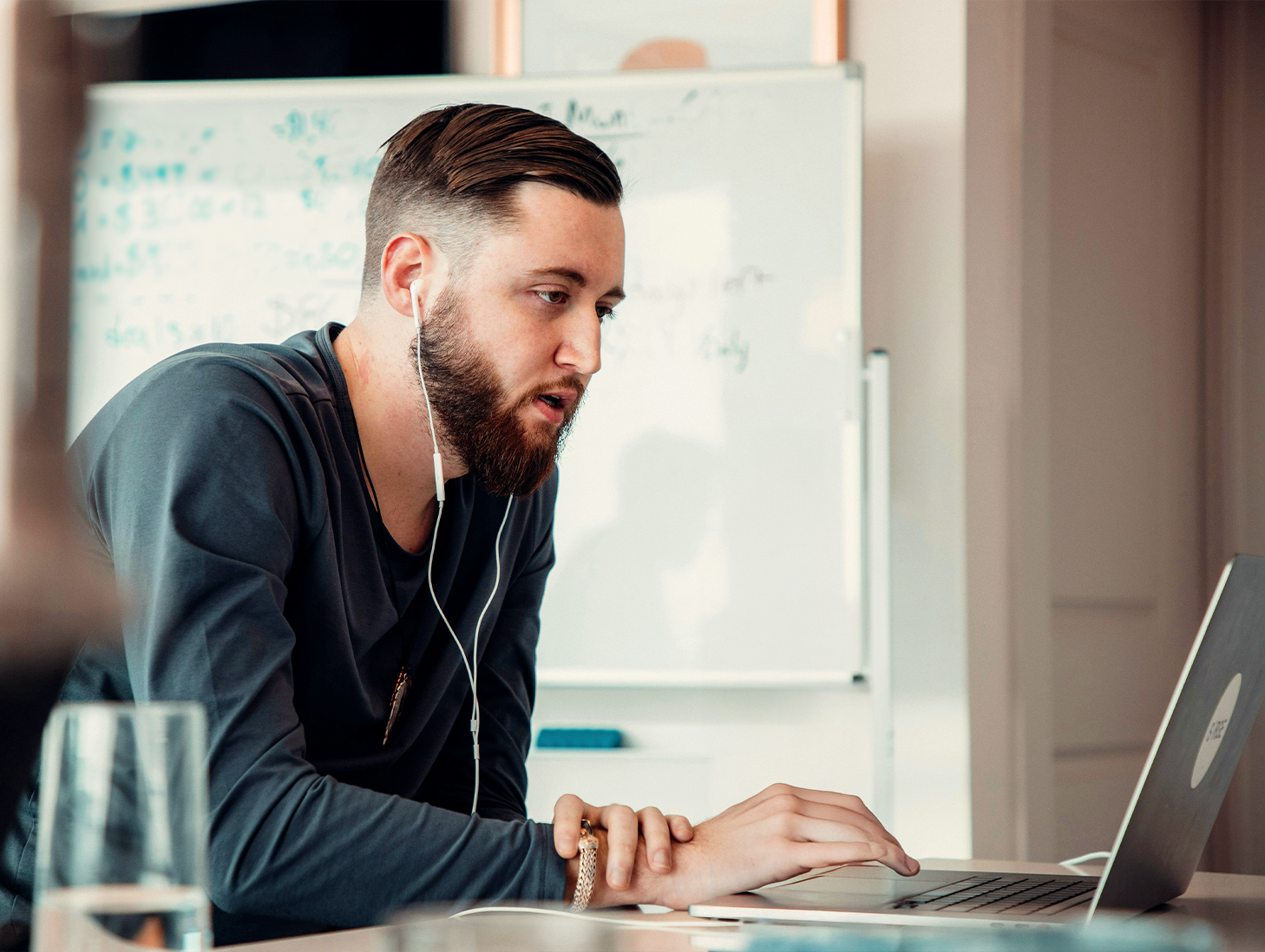 Man med hörlurar som arbetar på en bärbar dator vid ett skrivbord, whiteboard med skrift i bakgrunden - perfekt för att fånga den fokuserade studieatmosfären på yrkeshögskolan.