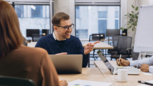 En man ler och gestikulerar medan han pratar med kollegor på ett kontorsmöte med bärbara datorer på bordet.