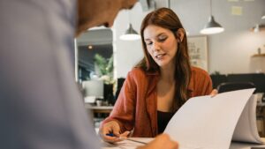 Kvinna i orange skjorta diskuterar dokument med en kollega vid ett skrivbord på ett modernt kontor.