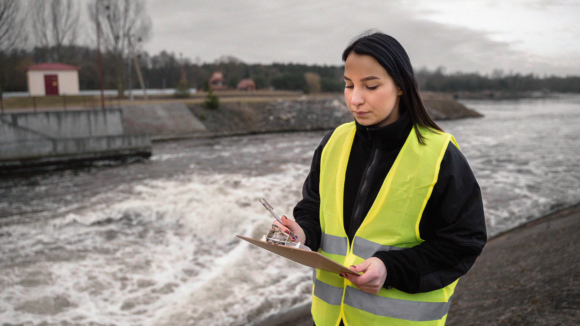 Kvinna i gul skyddsväst håller i en skrivplatta och observerar vattnet på en industrianläggning utomhus.