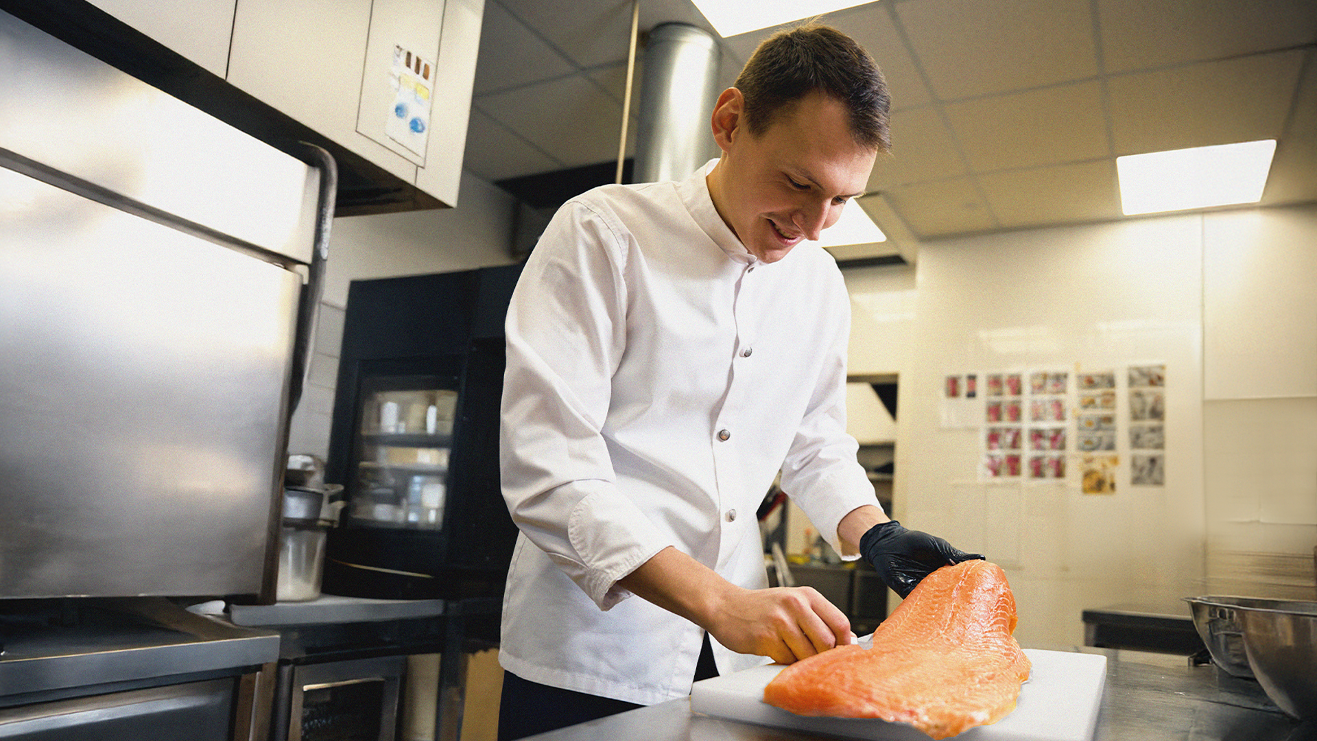 Kock i vit rock förbereder en stor laxfilé på en skärbräda i ett professionellt kök.