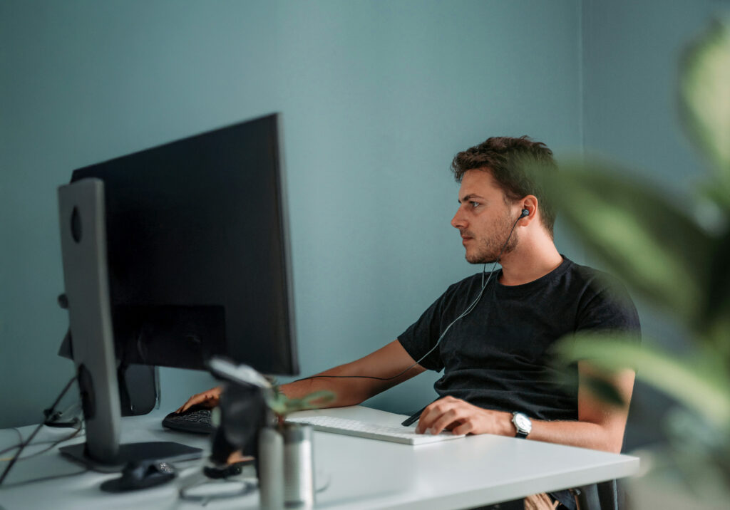 Man med hörlurar som arbetar vid ett datorbord mot en blågrön vägg med en växt i förgrunden - en perfekt bild av yrkeshögskolestudenternas fokuserade studiemiljö.