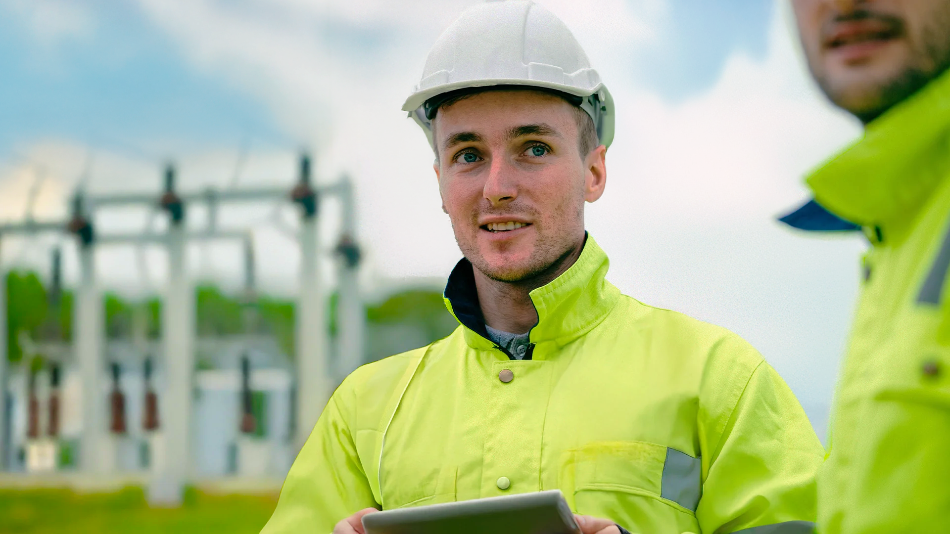 En Elkraftsingenjör i skyddshjälm och varseljacka håller i en surfplatta på ett industriområde utomhus.