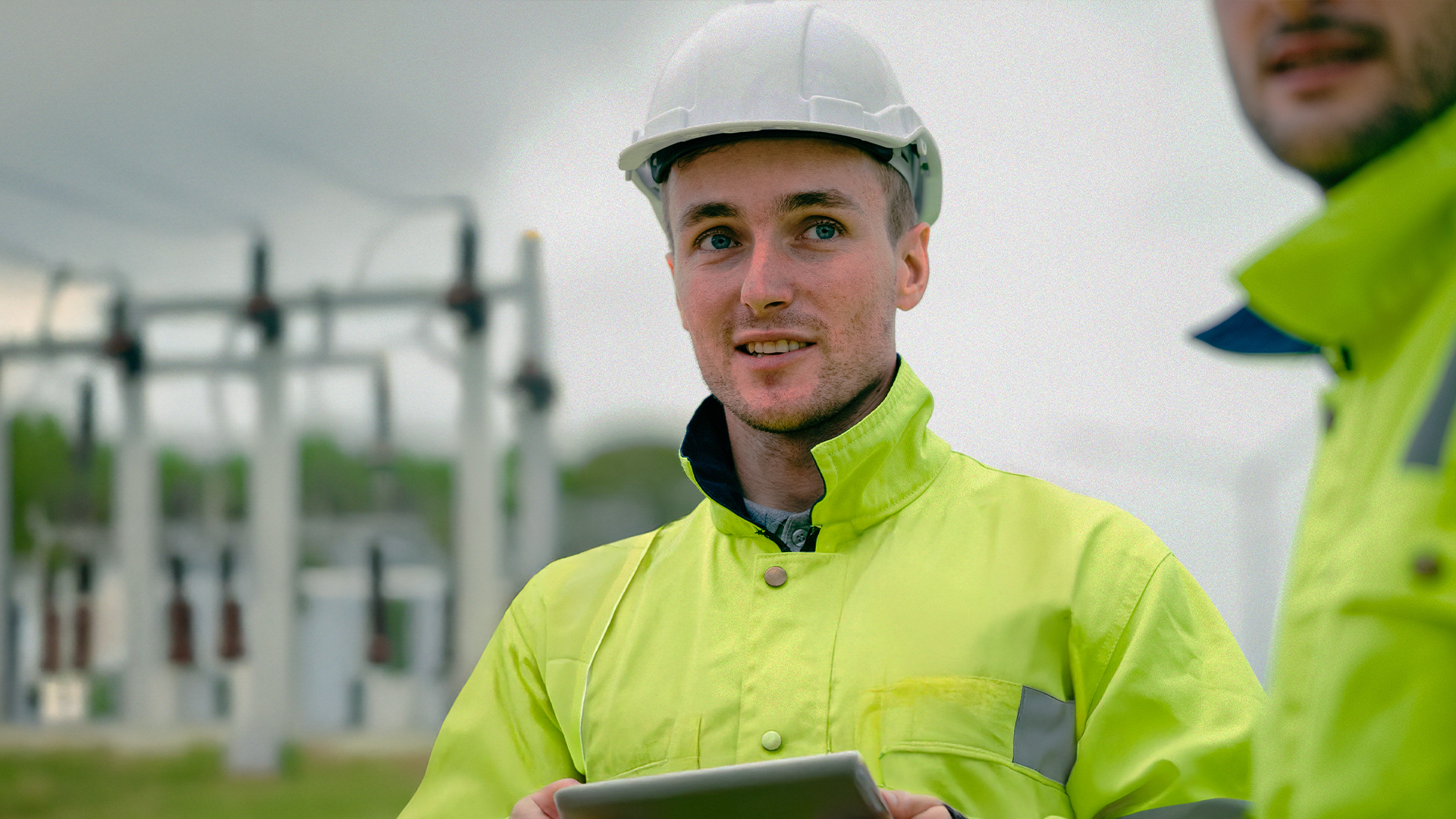 Man i gul skyddsjacka och skyddshjälm håller i en surfplatta på en industrianläggning utomhus.