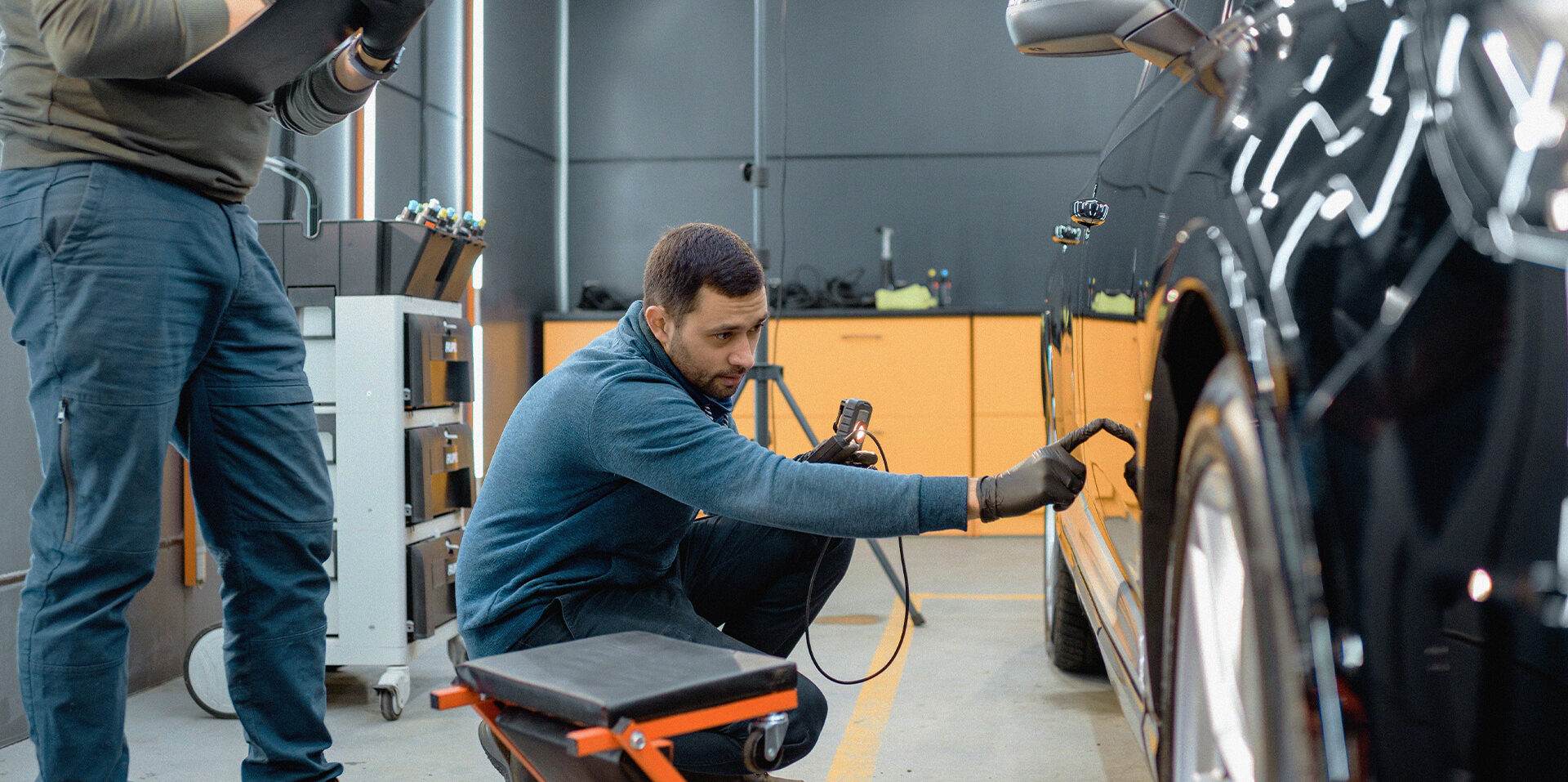 En man står på knä och inspekterar en bilsida med ett verktyg i ett garage; en annan person står i närheten.