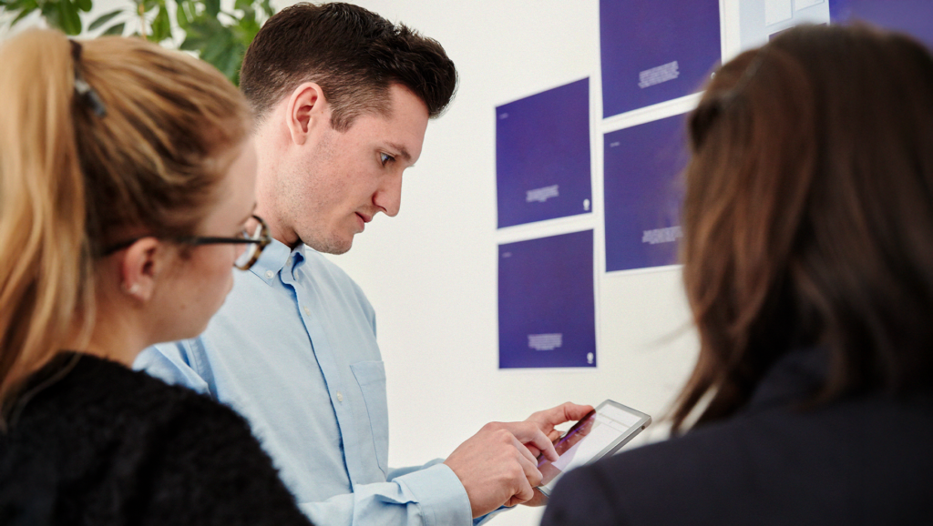 En man som använder en surfplatta förklarar något för två kvinnor framför en vägg med blå papper.