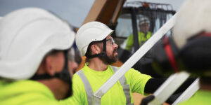 Byggnadsarbetare i skyddsutrustning och hjälm arbetar tillsammans på en byggarbetsplats med maskiner i bakgrunden.