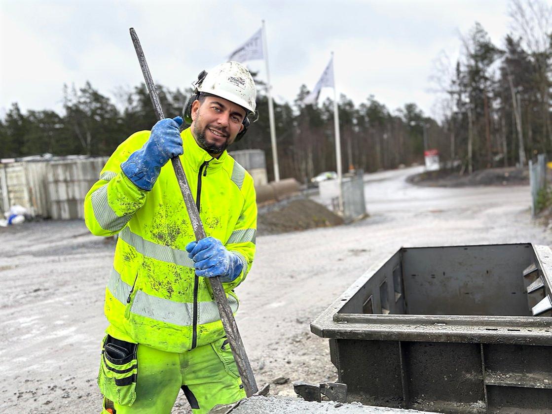 En leende byggnadsarbetare i neonjacka och hjälm står vid en sopcontainer med en metallstav i handen i utbildningen Betongingenjör.